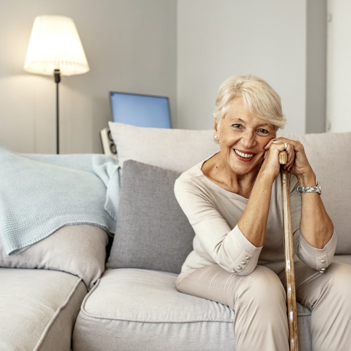 Photo of Retired short hair old woman with her walking stick at home looking at camera during the day while sitting on sofa. Senior gray hair women supporting on a walking cane at home.