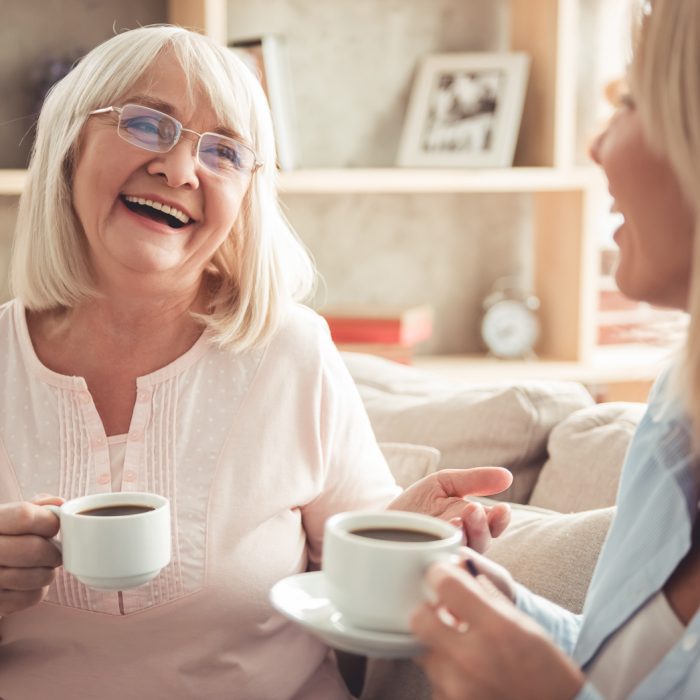 Beautiful mature mother and her adult daughter are drinking coffee, talking and smiling while sitting on couch at home