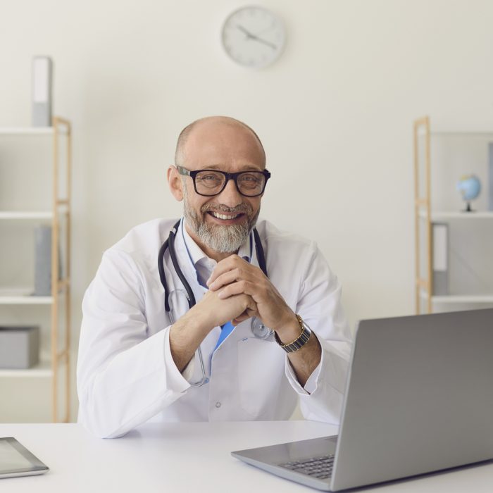 Mature doctor. A middle-aged confident doctor smiles while sitting at a table with a laptop at the workplace in the clinic office. Consultation of a medical professional in a hospital.